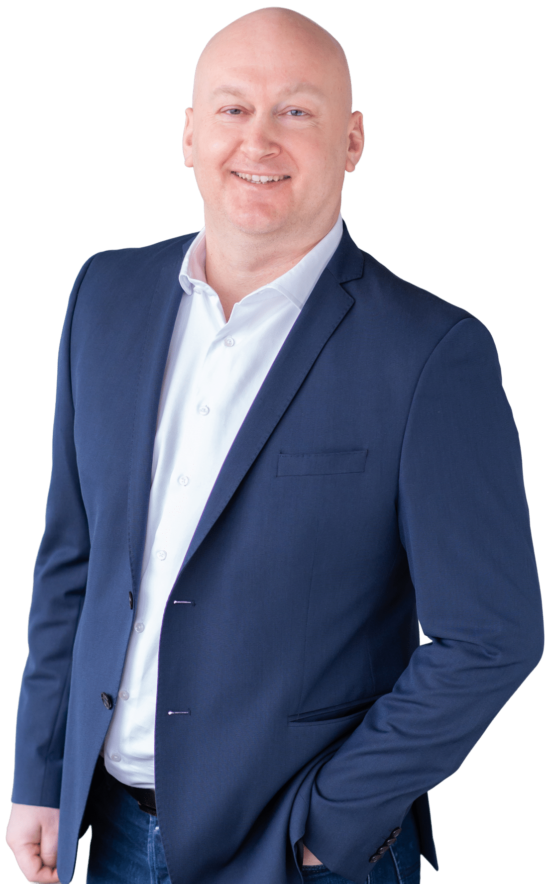 Ein glatzköpfiger Mann in einem marineblauen Blazer, weißem Hemd und Jeans steht lächelnd mit einer Hand in der Tasche vor einem schlichten weißen Hintergrund.