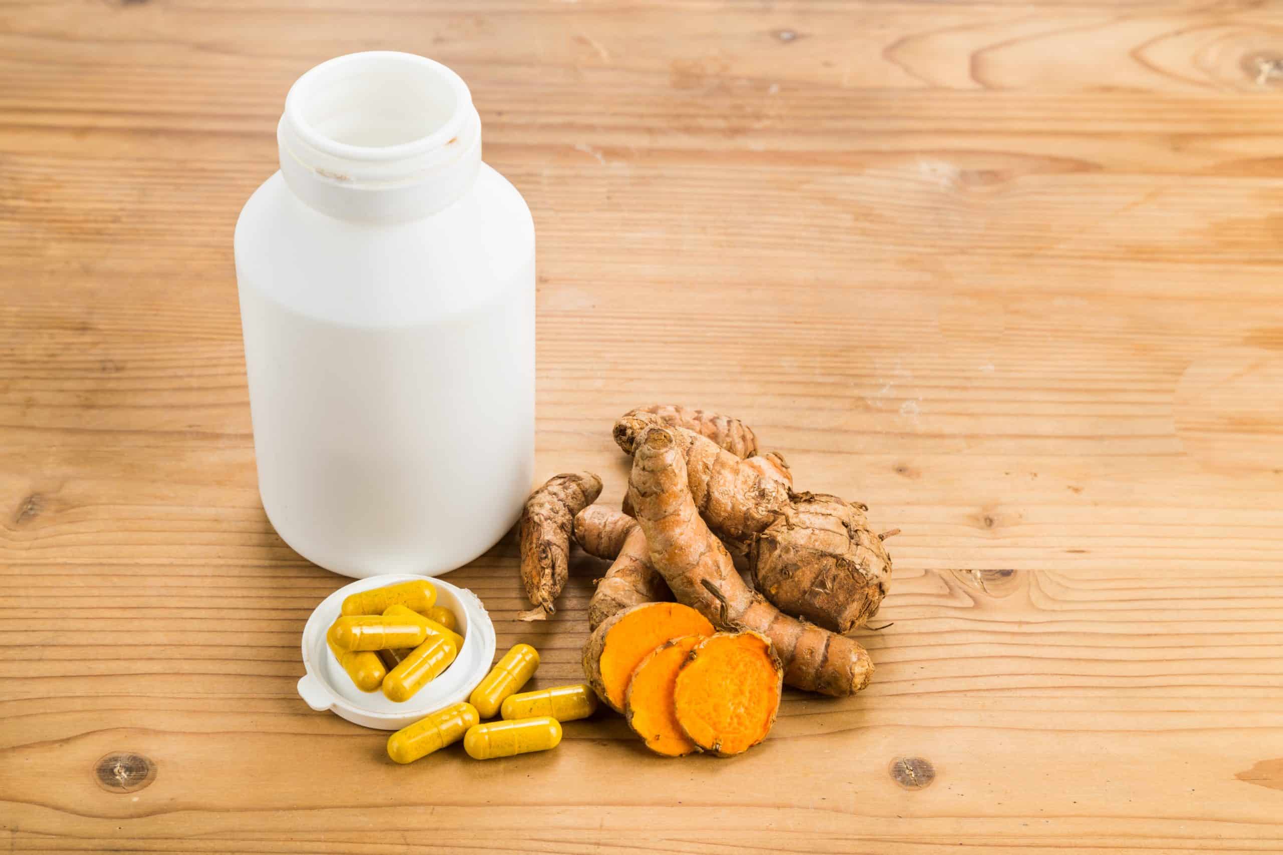 Ein weißes Pillenfläschchen mit gelben Kapseln auf dem Deckel liegt neben Kurkumawurzeln und -scheiben auf einer Holzfläche.