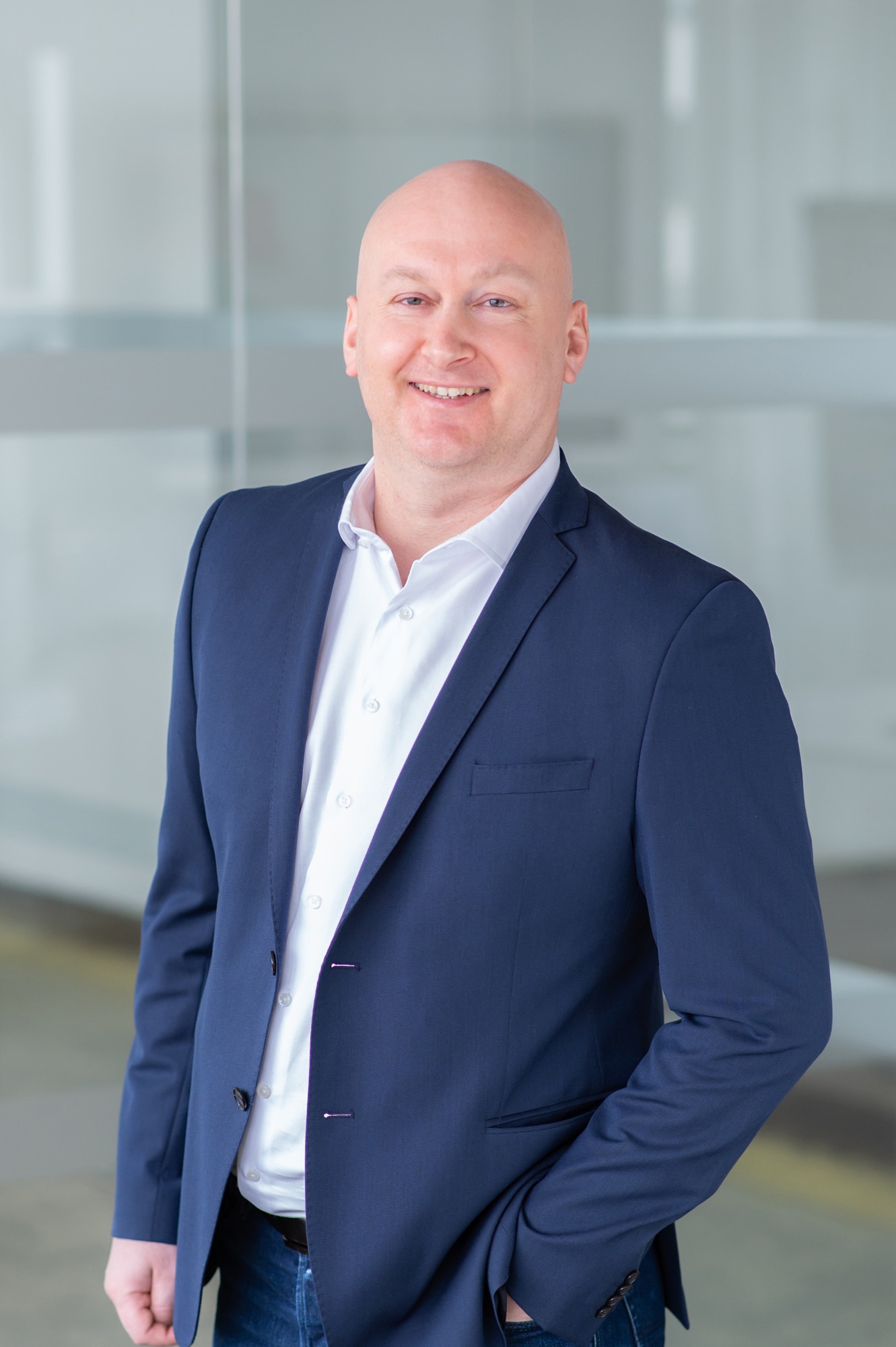 Ein lächelnder, glatzköpfiger Mann mit marineblauer Anzugsjacke und weißem Hemd steht mit einer Hand in der Tasche in einem hellen, modernen Büro mit Glaswänden.
