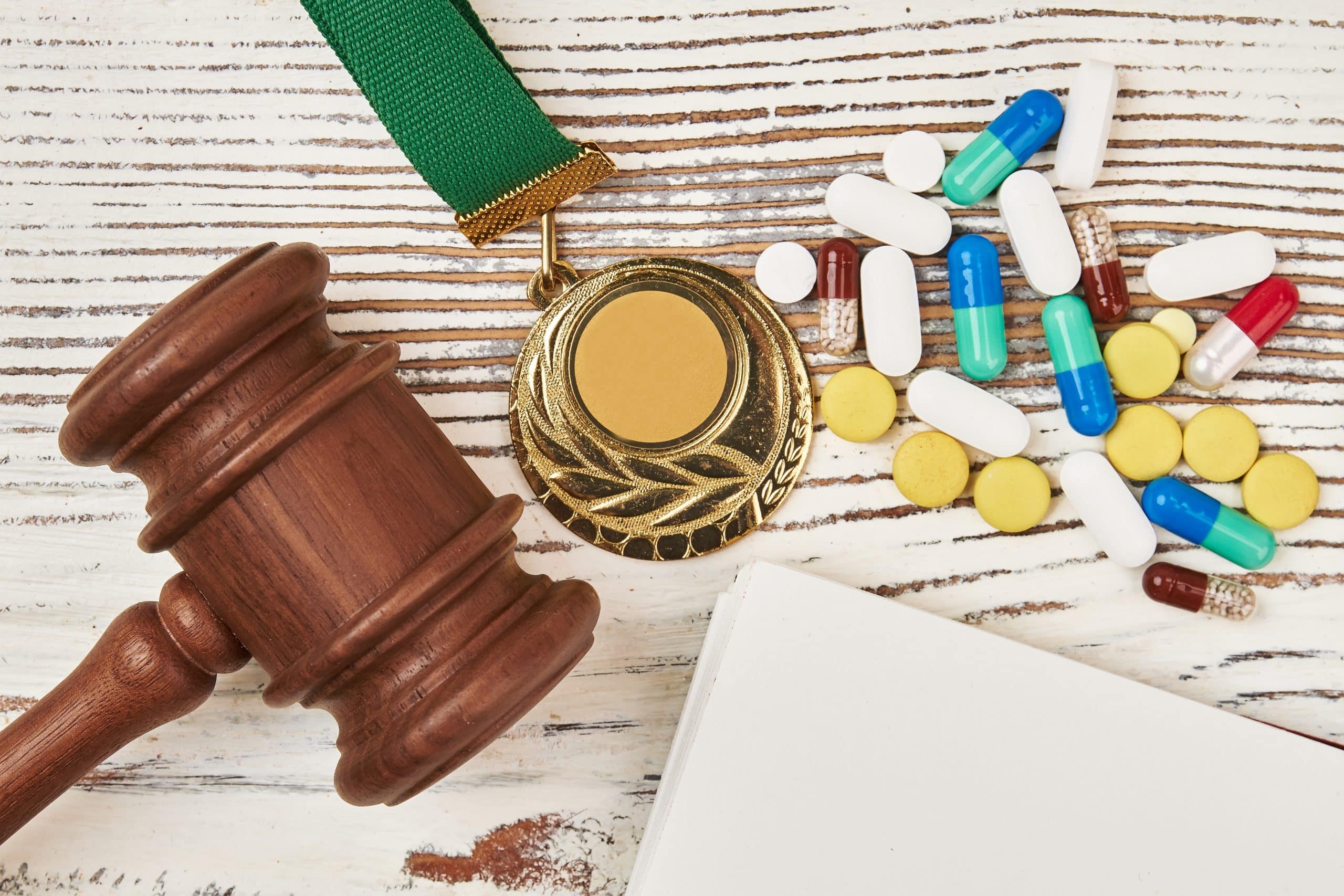Ein Richterhammer, eine Goldmedaille an einem grünen Band und verschiedene bunte Pillen sind auf einer weißen Holzfläche angeordnet.