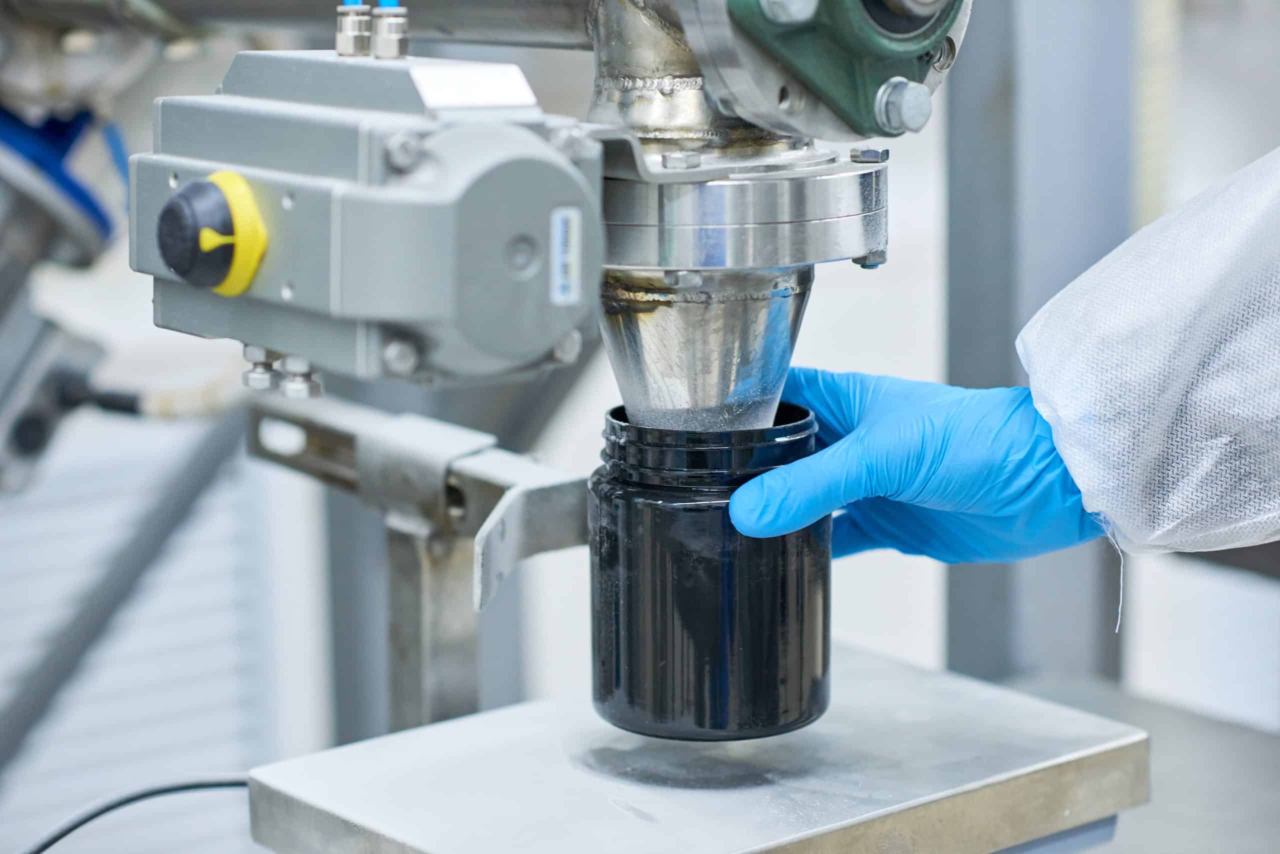 Eine Person, die einen blauen Handschuh trägt, hält ein schwarzes Gefäß unter einem Metallspender, wahrscheinlich in einem Labor oder einer Fabrik, um ein Produkt abzufüllen oder zu verpacken.