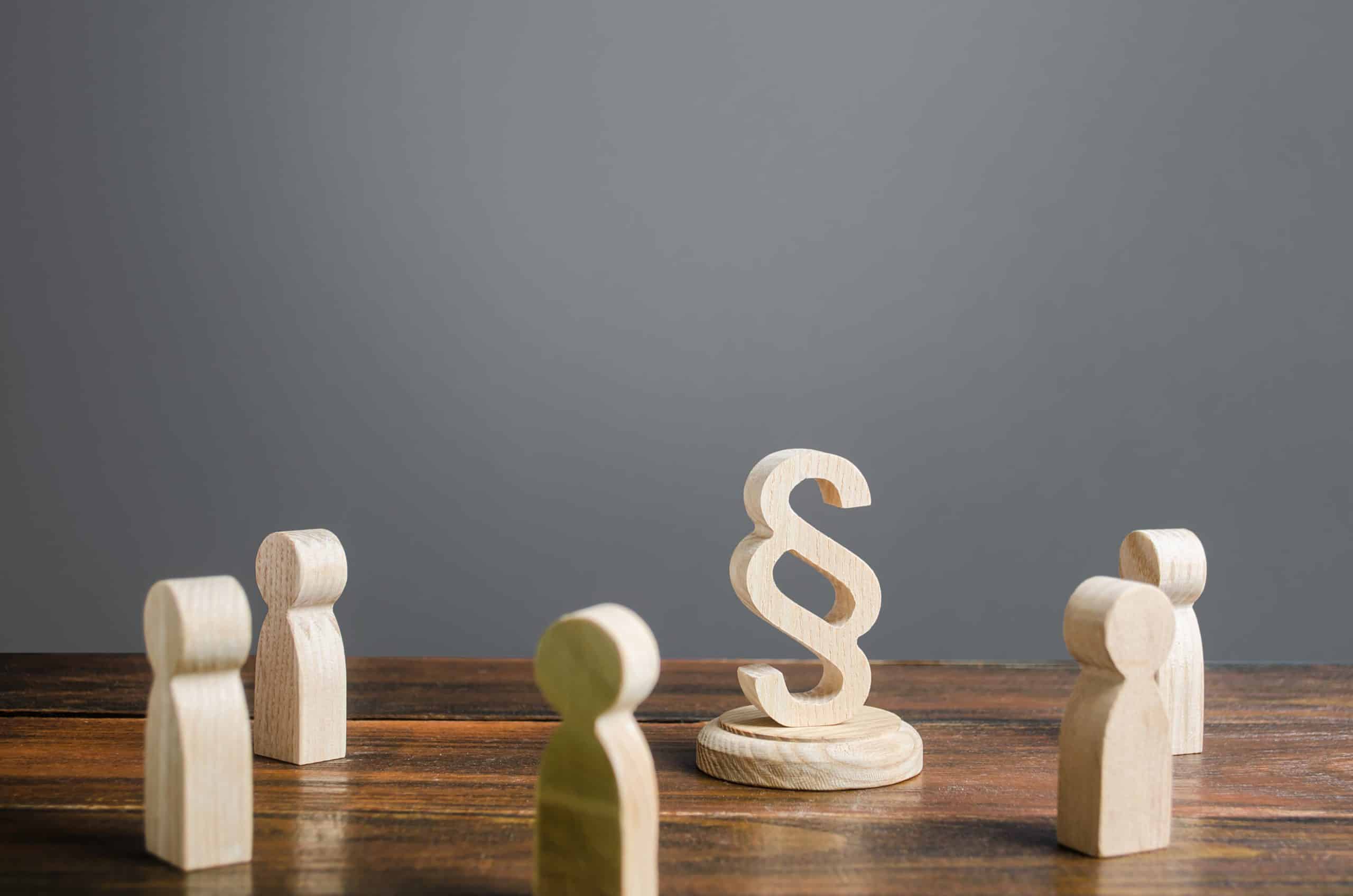 Vier hölzerne menschliche Figuren stehen um ein hölzernes Paragraphen-Symbol auf einem Holztisch, das Recht oder juristische Diskussionen symbolisiert, mit einem schlichten grauen Hintergrund.
