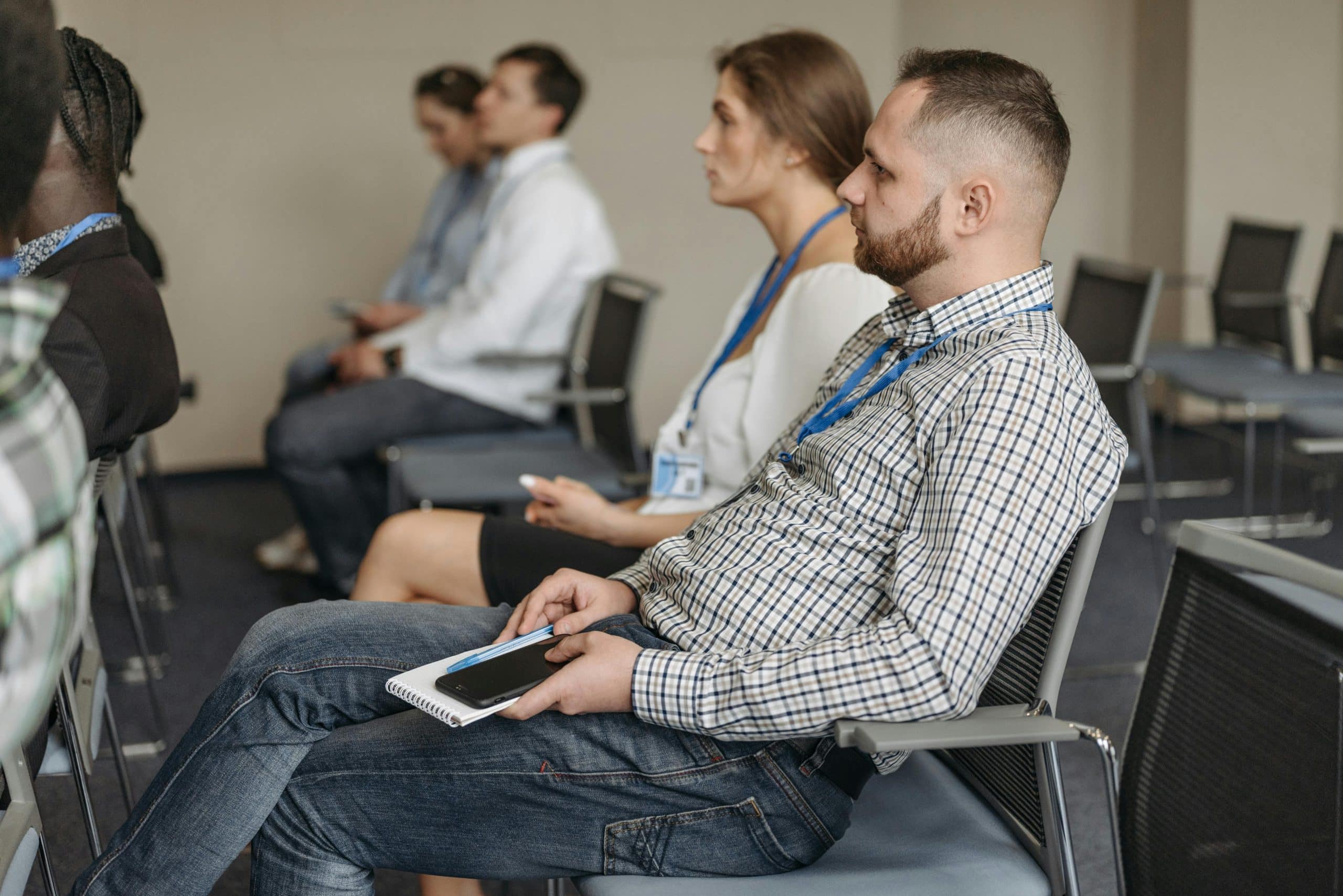 Eine Gruppe von Personen sitzt auf Stühlen in einem Konferenzraum und hört aufmerksam zu. Einige halten Notizbücher in der Hand und tragen blaue Lanyards mit Namensschildern. Der Fokus liegt auf einem Mann in einem karierten Hemd in der ersten Reihe.