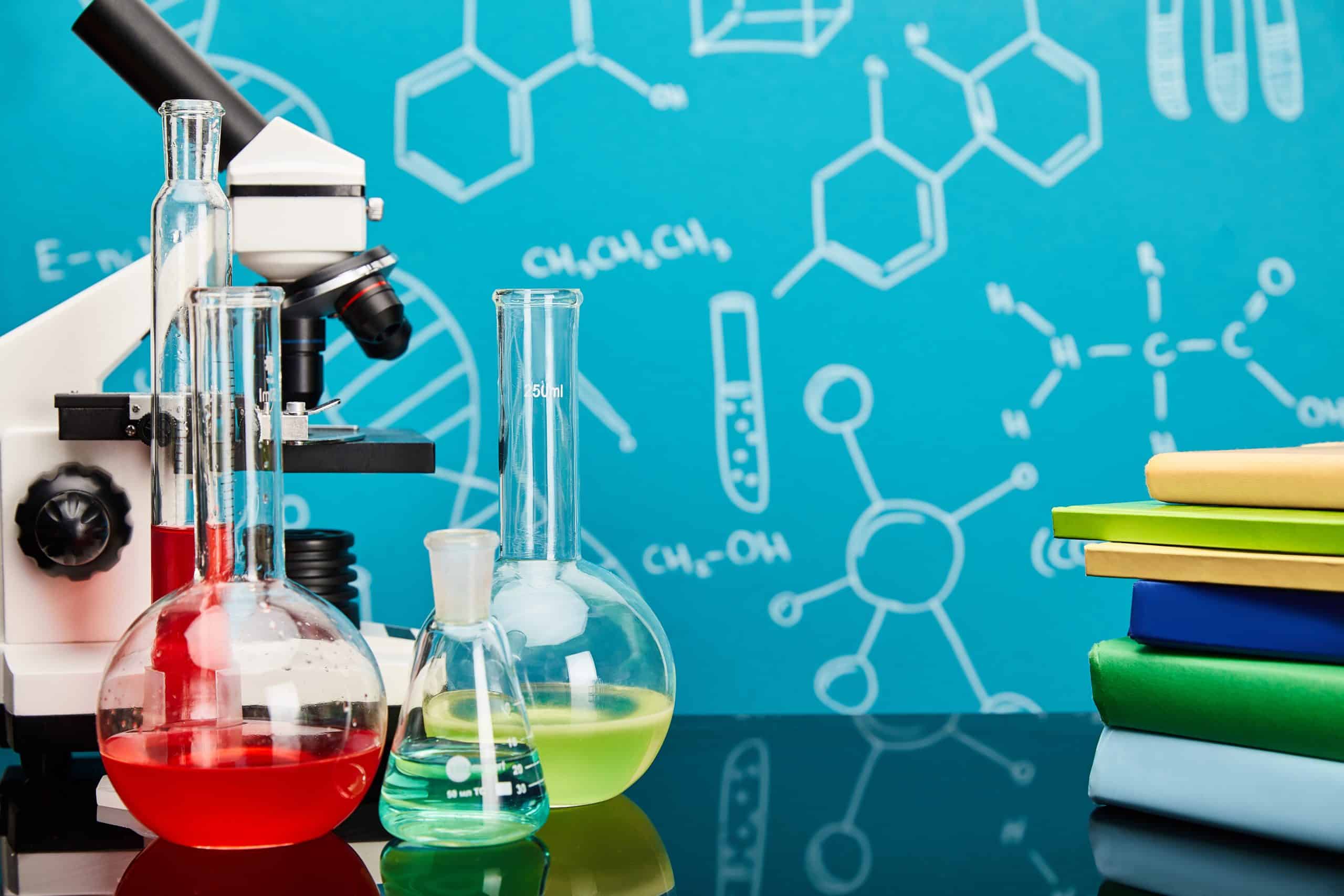 Ein Mikroskop und drei Laborglaskolben mit bunten Flüssigkeiten stehen auf einer schwarzen Fläche, daneben stapelweise Bücher und ein blauer Hintergrund mit weißen chemischen Formeln und Diagrammen.