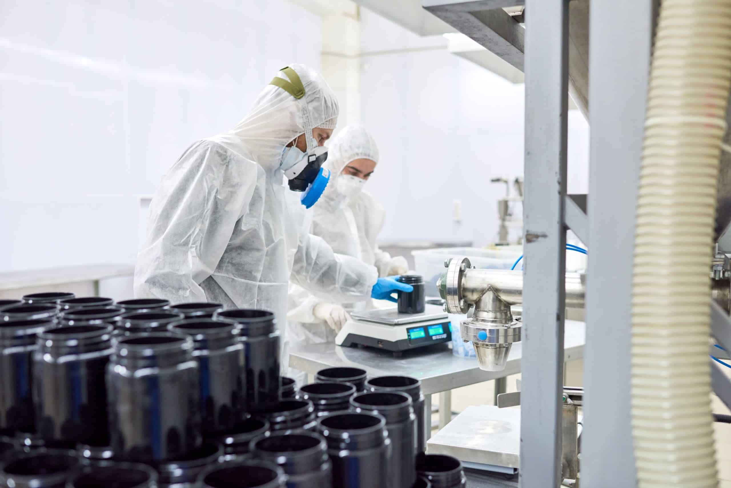 Zwei Personen in Schutzanzügen, Masken und Handschuhen arbeiten in einem Labor und stellen Behälter auf eine digitale Waage. Schwarze Plastikgefäße sind auf einem Tisch im Vordergrund gestapelt. Im Hintergrund ist Industrieausrüstung zu sehen.