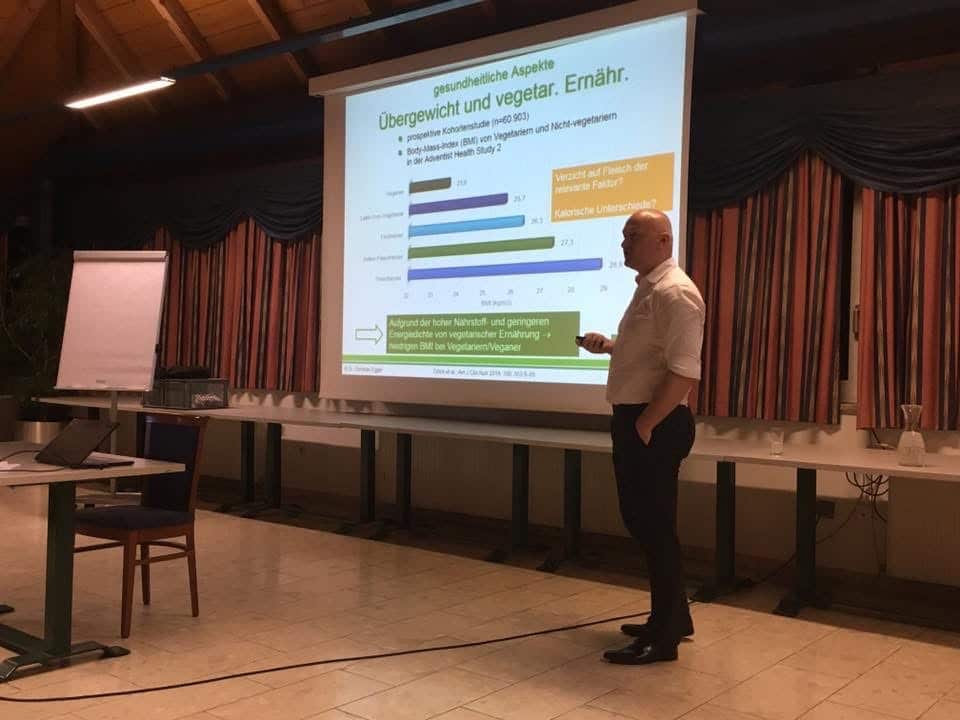 Ein Mann steht in der Nähe eines Projektors und hält einen Vortrag in deutscher Sprache über Übergewicht und vegetarische Ernährung, wobei er Grafiken und Texte anzeigt. Im Raum sind ein Flipchart und leere Stühle zu sehen.